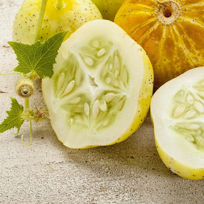 A photo showing fresh lemon cucumbers, with one cut in half to display the seeds, alongside a leaf and part of the fruit's skin, on a stone surface.