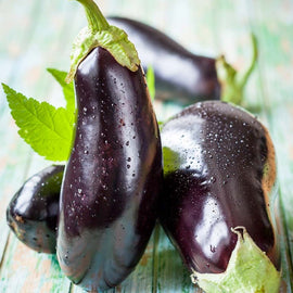 Several fresh eggplants with deep purple skin and green leaves, droplets of water visible on the surface, arranged on a wooden surface.
