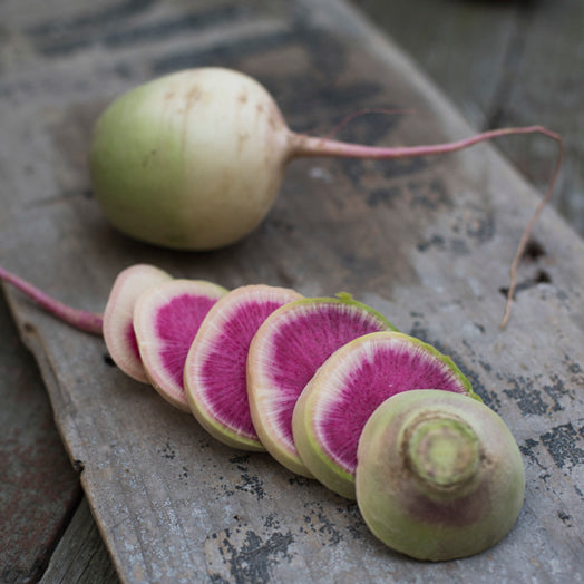 A whole watermelon radish with a green skin and a pink watermelon-like flesh, cut into thin slices on a wooden surface.