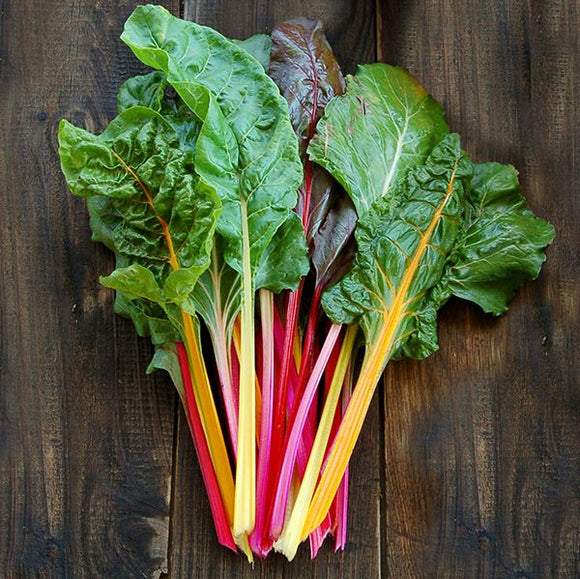 A bunch of Swiss Chard with colorful stems in red, yellow, and pink hues, laid out on a wooden surface.